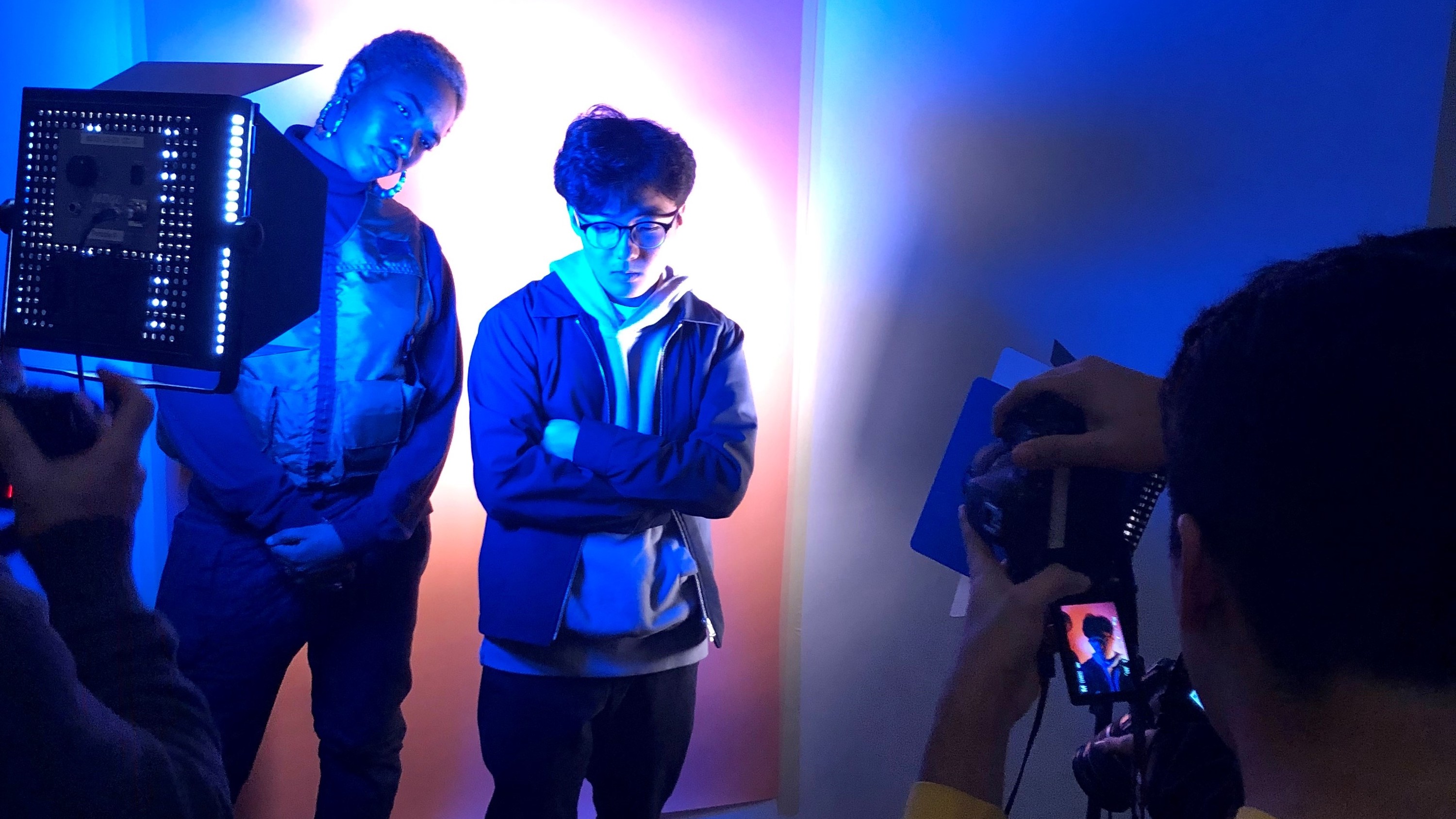 Two teens stand in a serious pose with bright white and blue lights on them while  someone off the frame takes a photo of them