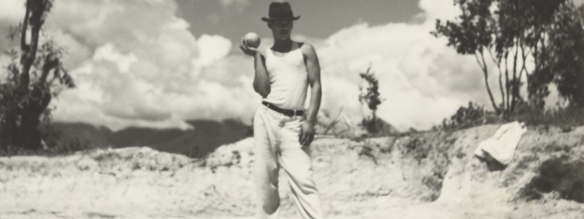 In front of mountains and a bright cloud filled sky, a man dressed in white pants, white t-shirt, and a dark brimmed hat, stands prepared to throw the ball he's holding up in his right palm.